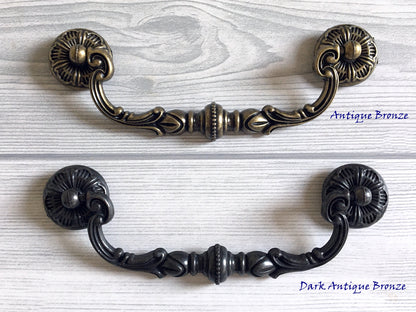 Two cabinet swing bail handles, one in antique bronze and one in dark antique bronze, on a light wooden surface.